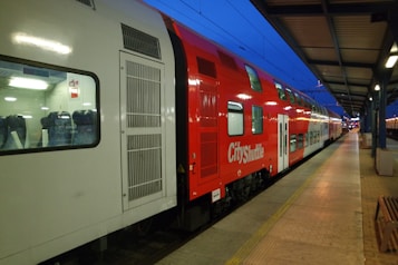 A brightly colored red and white double-decker train labeled 'City Shuttle' is parked at a platform in a train station. The train features large windows with visible seats inside. The platform is empty, and the overhead roof stretches along the platform, with lights illuminating the scene. The sky is a deep blue, suggesting it is either dawn or dusk.