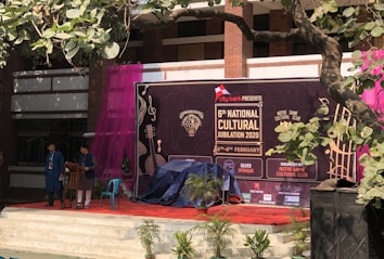 A stage setup for the 6th National Cultural Jubilation 2020 event, organized by the Notre Dame Cultural Club. There are two people standing on stage near a podium, with a large banner in the background featuring musical instruments and event details. The area is decorated with potted plants, and there is a purple curtain on the side. A tree branch with green leaves partially covers the view.