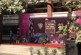 A stage setup for the 6th National Cultural Jubilation 2020 event, organized by the Notre Dame Cultural Club. There are two people standing on stage near a podium, with a large banner in the background featuring musical instruments and event details. The area is decorated with potted plants, and there is a purple curtain on the side. A tree branch with green leaves partially covers the view.