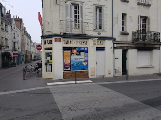 A street corner featuring an old European-style building with signs indicating a tobacco shop, press, and bar. The facade displays advertisements and a closed door. The street is quiet, with bicycles and tables outside. The surrounding buildings have classic architecture with shutters and balconies.