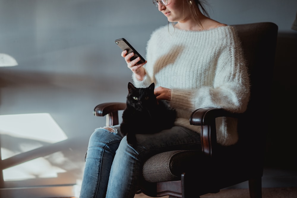 woman in white sweater and blue denim jeans sitting on black leather armchair holding silver iphone