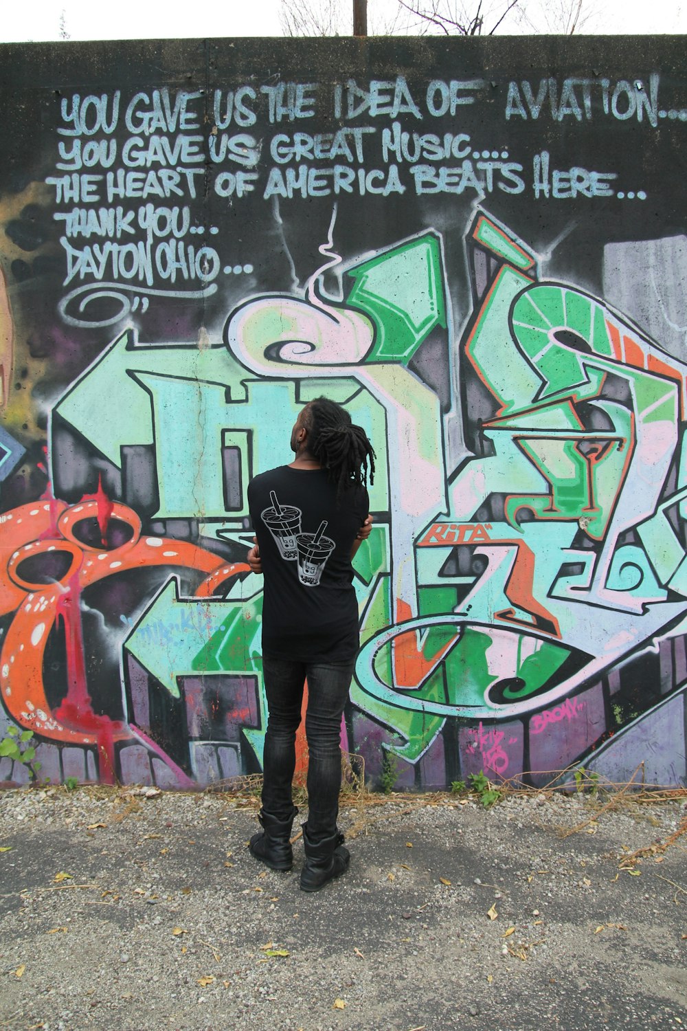 mulher na camisa preta de manga comprida e calças pretas em pé ao lado da parede do grafite durante o dia