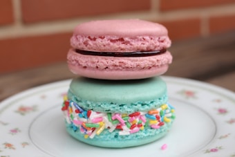 A pair of macarons stacked on top of each other are placed on a floral-patterned plate. The top macaron is pink with a dark filling, while the bottom macaron is light blue with colorful sprinkles around its filling. The background includes a blurred red brick wall.