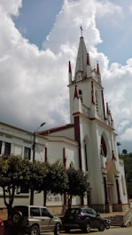 A tall, white church building with pointed spires and red accents stands prominently under a partially cloudy sky. The church features arched windows and a large entrance. Several cars are parked in front of it, near a row of trees lining the street.