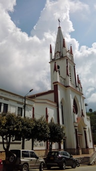 A tall, white church building with pointed spires and red accents stands prominently under a partially cloudy sky. The church features arched windows and a large entrance. Several cars are parked in front of it, near a row of trees lining the street.