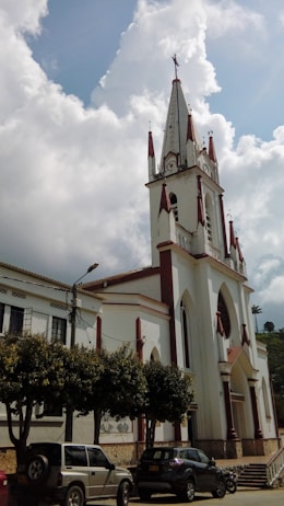A tall, white church building with pointed spires and red accents stands prominently under a partially cloudy sky. The church features arched windows and a large entrance. Several cars are parked in front of it, near a row of trees lining the street.