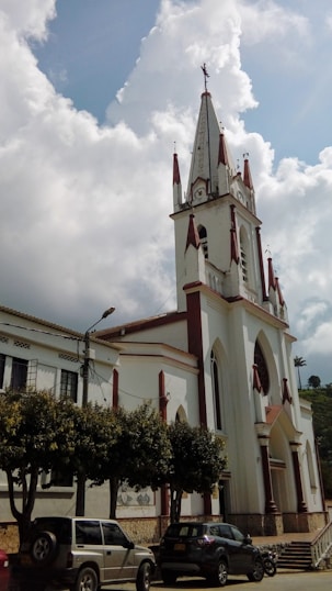 A tall, white church building with pointed spires and red accents stands prominently under a partially cloudy sky. The church features arched windows and a large entrance. Several cars are parked in front of it, near a row of trees lining the street.