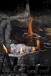 A piece of wood is burning inside a fireplace. Flames rise towards the dark, sooty background, while the wood is charred and partially white from ash.