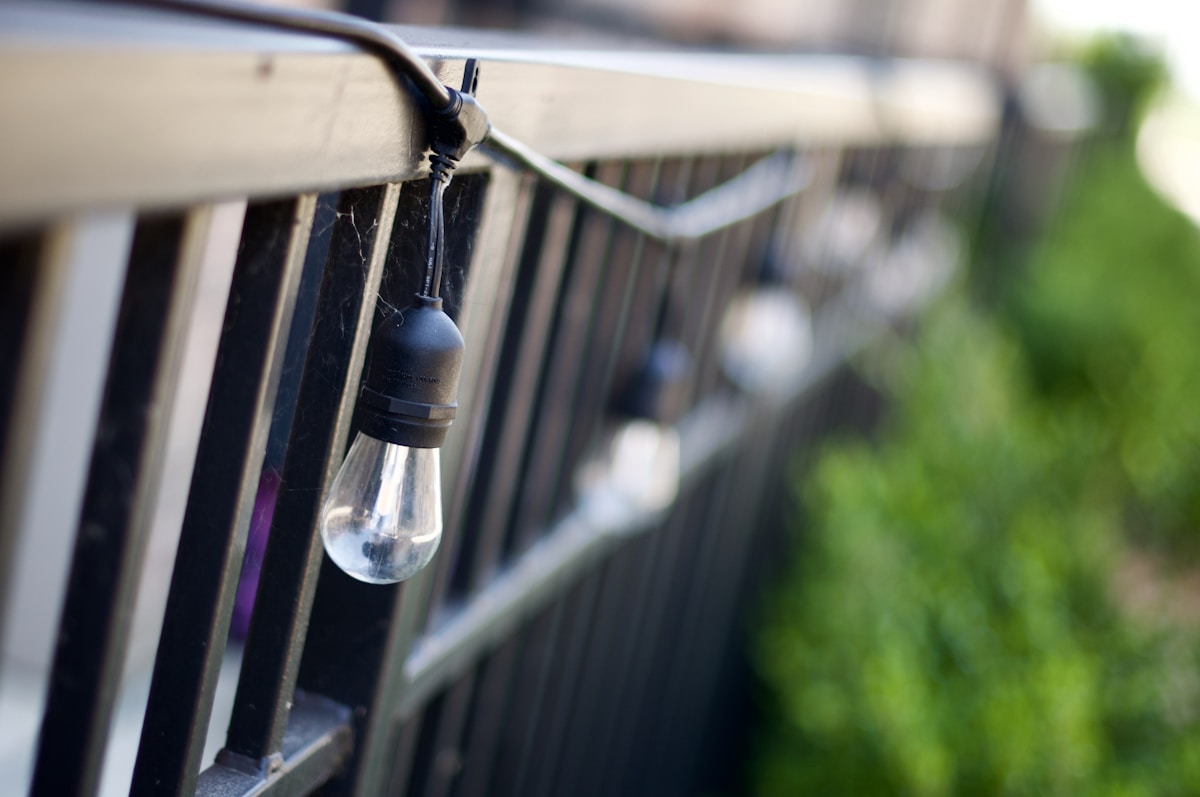Edison style string light bulbs hanging from the railing of an apartment balcony with green garden plants visible in the background