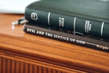 A close-up of two books stacked on a wooden surface. The top book is a green Holy Bible with the title and Christian Standard Bible label in white text. Below it, a dark brown book titled 'Evil And The Justice Of God' is visible.