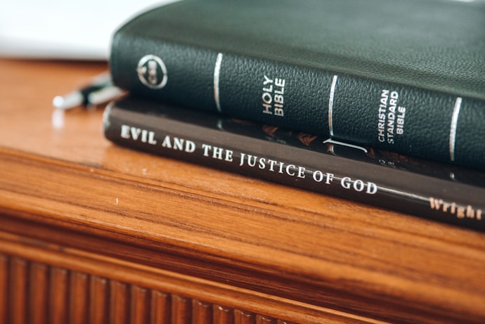 A close-up of two books stacked on a wooden surface. The top book is a green Holy Bible with the title and Christian Standard Bible label in white text. Below it, a dark brown book titled 'Evil And The Justice Of God' is visible.