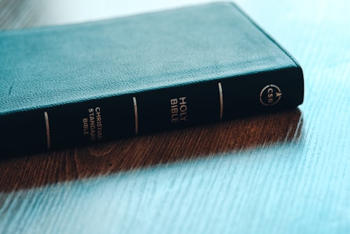 A hardcover Christian Standard Bible lies on a wooden table. The spine of the book is visible, showcasing the title 'Holy Bible' along with the abbreviation 'CSB'. The texture of the book cover appears to be leather, and the table has a polished wood grain finish.