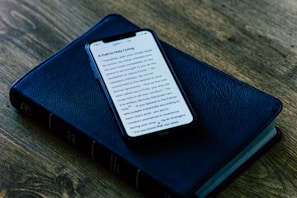 A smartphone displaying text on its screen is placed on top of a closed, black, leather-bound book, which appears to be a Bible. The setup is on a wooden surface, giving a rustic and serene ambiance.