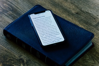 A smartphone displaying text on its screen is placed on top of a closed, black, leather-bound book, which appears to be a Bible. The setup is on a wooden surface, giving a rustic and serene ambiance.