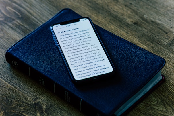 A smartphone displaying text on its screen is placed on top of a closed, black, leather-bound book, which appears to be a Bible. The setup is on a wooden surface, giving a rustic and serene ambiance.