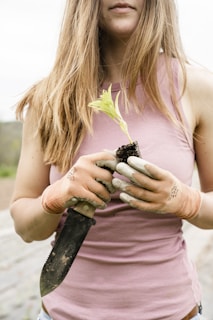 A woman with long, brown hair holds a small plant seedling in one hand and a gardening trowel in the other. She is wearing a sleeveless pink top and orange gardening gloves. The background is blurred, suggesting an outdoor setting.