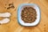 A bowl filled with dry dog food sits on a wooden floor. Next to it are a pair of dog paws and some scattered kibble pieces.