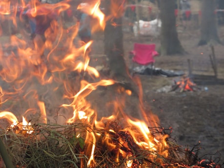 Flames and smoke rise from a pile of burning branches and twigs in a wooded area. In the background, a red chair and a smaller campfire can be seen. The scene takes place outdoors, likely in a forest.