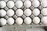 A close-up view of a full carton with white eggs arranged in neat rows. The eggs are placed in a cardboard egg tray, showing a slightly rough texture and grey-brown color of the cardboard.
