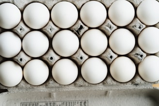 A close-up view of a full carton with white eggs arranged in neat rows. The eggs are placed in a cardboard egg tray, showing a slightly rough texture and grey-brown color of the cardboard.