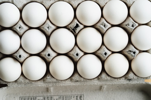 A close-up view of a full carton with white eggs arranged in neat rows. The eggs are placed in a cardboard egg tray, showing a slightly rough texture and grey-brown color of the cardboard.