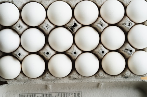 A close-up view of a full carton with white eggs arranged in neat rows. The eggs are placed in a cardboard egg tray, showing a slightly rough texture and grey-brown color of the cardboard.