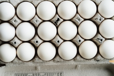 A close-up view of a full carton with white eggs arranged in neat rows. The eggs are placed in a cardboard egg tray, showing a slightly rough texture and grey-brown color of the cardboard.