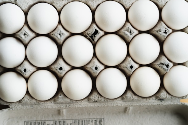A close-up view of a full carton with white eggs arranged in neat rows. The eggs are placed in a cardboard egg tray, showing a slightly rough texture and grey-brown color of the cardboard.