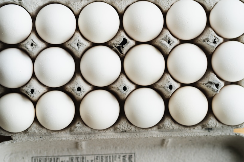 A close-up view of a full carton with white eggs arranged in neat rows. The eggs are placed in a cardboard egg tray, showing a slightly rough texture and grey-brown color of the cardboard.