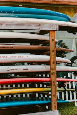 A stack of surfboards is neatly arranged on a wooden rack. The rack is situated outdoors, in front of a picket fence, with a small glimpse of greenery in the background. The surfboards vary in colors including blue, white, red, yellow, and some show signs of wear and tear.
