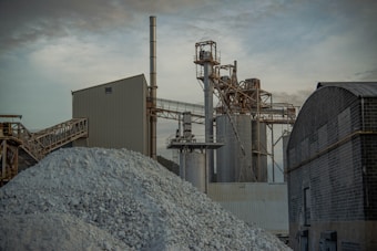 An industrial facility with multiple large structures and smokestacks is evident. Piles of rock or gravel are in the foreground, with various metal frameworks and a conveyor system. The sky is overcast, adding a somber tone to the scene.