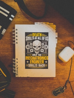 A notebook with a spiral binding is on a wooden surface. The cover features a design with a skull and text in bold yellow and white letters that reads, 'Death Smiles at All of Us, Only the Mechatronics Engineer Smiles Back.' Nearby, there is a white rectangular object, a keychain with a metallic tag, and a bag or pouch with a striped pattern.