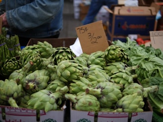 A market stall displaying a large pile of fresh artichokes with a handwritten sign indicating the price. Other green vegetables, such as asparagus, are visible nearby. In the background, cardboard boxes and a person wearing a denim jacket can be seen.