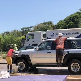Two people are engaged in washing a silver SUV. One is standing on the ground, cleaning the front of the vehicle, and the other is reaching up to clean the side. There is a bucket of soapy water on the ground being used for the task. The background shows a large truck and some greenery under a clear blue sky.