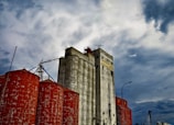 A large industrial grain silo complex with weathered white silos and rust-colored cylindrical tanks, set against a dramatic, cloudy sky.