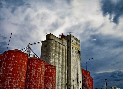 A large industrial grain silo complex with weathered white silos and rust-colored cylindrical tanks, set against a dramatic, cloudy sky.