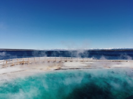 A geothermal hot spring with vibrant turquoise water emits steam under a clear blue sky. A wooden boardwalk encircles the spring, providing a pathway for visitors. In the background, a large body of water stretches out, leading to a distant line of snow-capped mountains.