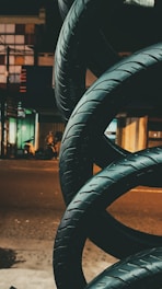 A stack of car tires is arranged in an artistic spiral on the side of a street at night. Buildings with various light sources are visible in the background, giving a slightly industrial and urban atmosphere.