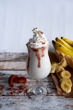 A glass filled with a creamy banana milkshake topped with whipped cream and sprinkled with small red bits, sitting on a rustic wooden surface. To the side, there is a cut banana and a bunch of whole bananas in a basket.