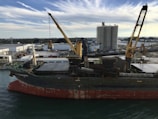 A large cargo ship is docked at an industrial port. The ship has a visible rusted hull and onboard cranes for loading and unloading. In the background, there are industrial buildings, storage tanks, and machinery. The sky is partly cloudy, with streaks of white clouds against a blue backdrop.