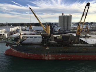 A large cargo ship is docked at an industrial port. The ship has a visible rusted hull and onboard cranes for loading and unloading. In the background, there are industrial buildings, storage tanks, and machinery. The sky is partly cloudy, with streaks of white clouds against a blue backdrop.