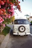 A vintage white van is parked on the side of a road near a sidewalk lined with vibrant pink flowers. The van has a spare tire mounted on the front grille and is surrounded by greenery and residential buildings in the background. A Californian license plate is visible at the front.