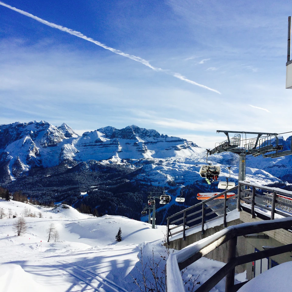 The image depicts a snowy mountain landscape with a ski lift ascending towards the top of the mountain, carrying skiers and snowboarders to the top. The sky is clear blue with a trail left by a plane flying overhead. In the foreground, there are trees dotting the landscape, adding depth and texture to the scene.
