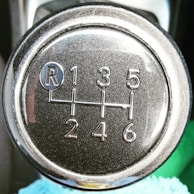 A close-up view of a metallic gear shift knob displaying gear numbers and the letter 'R' for reverse. The surface has a textured metallic sheen, and light reflects off its polished edges.