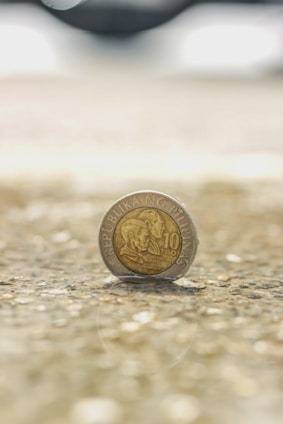 A Filipino ten peso coin is standing on its edge on a textured surface. The background is blurred, focusing the attention on the coin which features the inscription 'Republika ng Pilipinas' and portraits of two individuals.