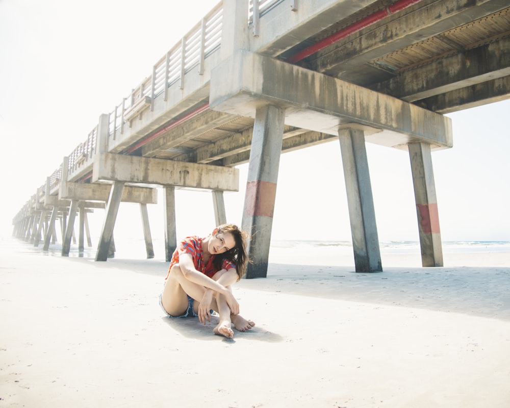 woman in red shirt sitting on white sand under gray concrete bridge during daytime