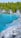 A stunning natural landscape featuring clear turquoise water surrounded by rugged rocky cliffs. Dense green trees form a lush backdrop, creating a tranquil and serene environment. The rocky shoreline presents a natural, unrefined beauty, with scattered patches of people exploring the area.