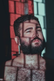 A bearded man is illuminated by natural light, casting deep shadows with a grid-like pattern across his face and chest. His expression is contemplative as he looks upward, and he is positioned against a partially lit red and dark backdrop.