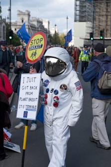 A person in a spacesuit is participating in a protest, surrounded by a crowd holding EU flags. The person holds a sign reading 'I just got back from planet Brexit. It is terrible' and a circular sign with 'Stop Brexit'. The scene is set on a city street with buildings and police in the background.