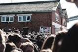 A large crowd of people gathered outdoors, many holding cameras or their phones aloft, appears to be at some sort of public event or protest. Prominently displayed in the center is a sign with the words 'ACT NOW' against a stylized background featuring a profile image. In the background, there is a red brick building with white window frames and a corrugated roof, as well as a partial view of a bridge.